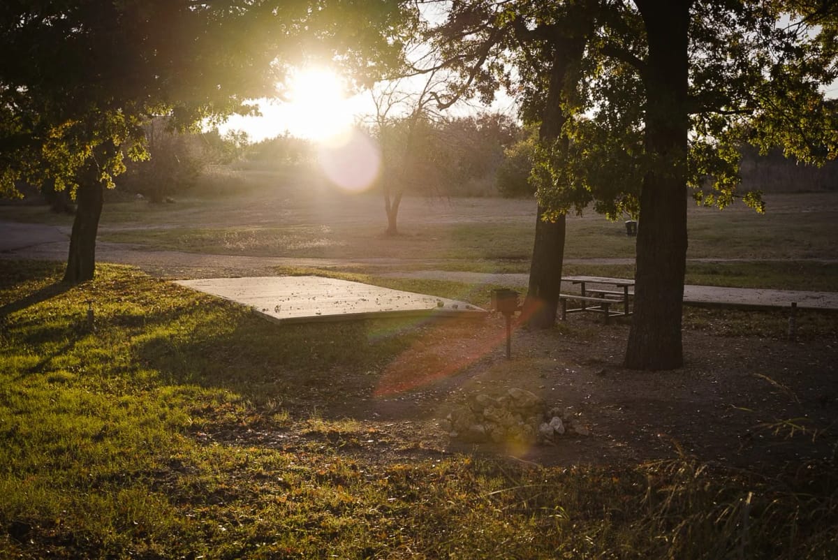 A park scene at sunset with trees, a picnic table, and a small fire pit, sunlight filtering through the trees.